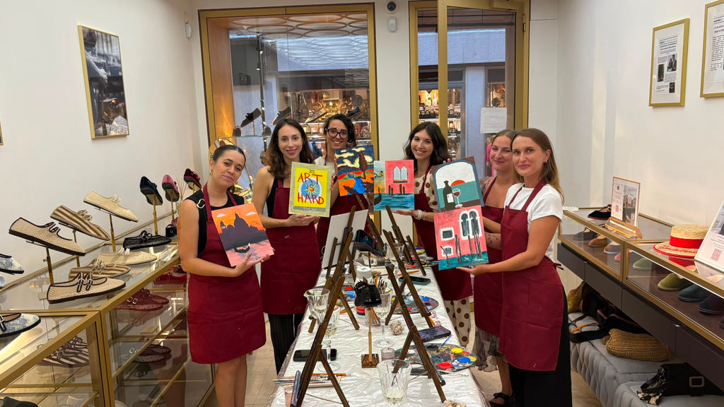 Five women wearing red aprons stand in a shop holding colorful painted canvases, with an easel in the center holding a painting. Shoe displays and framed wall art